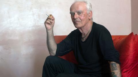Der Künstler Jürgen Klauke sitzt dunkel gekleidet auf einem roten Sofa. In der rechten Hand hält er eine Zigarette, seine linke, tätowierte Hand stützt sich auf dem Sofa ab. Um den Hals trägt er eine dicke Silberkette.