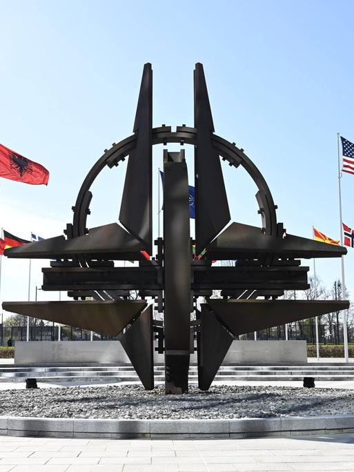Vor dem NATO-Hauptquartier flattern die Fahnen der Mitgliedsstaaten im Wind, sie sind kreisförmig um das NATO-Symbol aufgestellt.