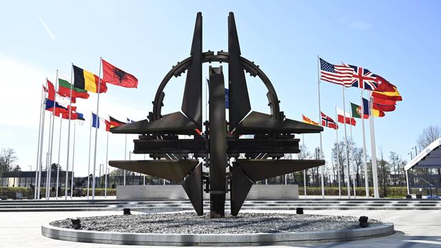Vor dem NATO-Hauptquartier flattern die Fahnen der Mitgliedsstaaten im Wind, sie sind kreisförmig um das NATO-Symbol aufgestellt.