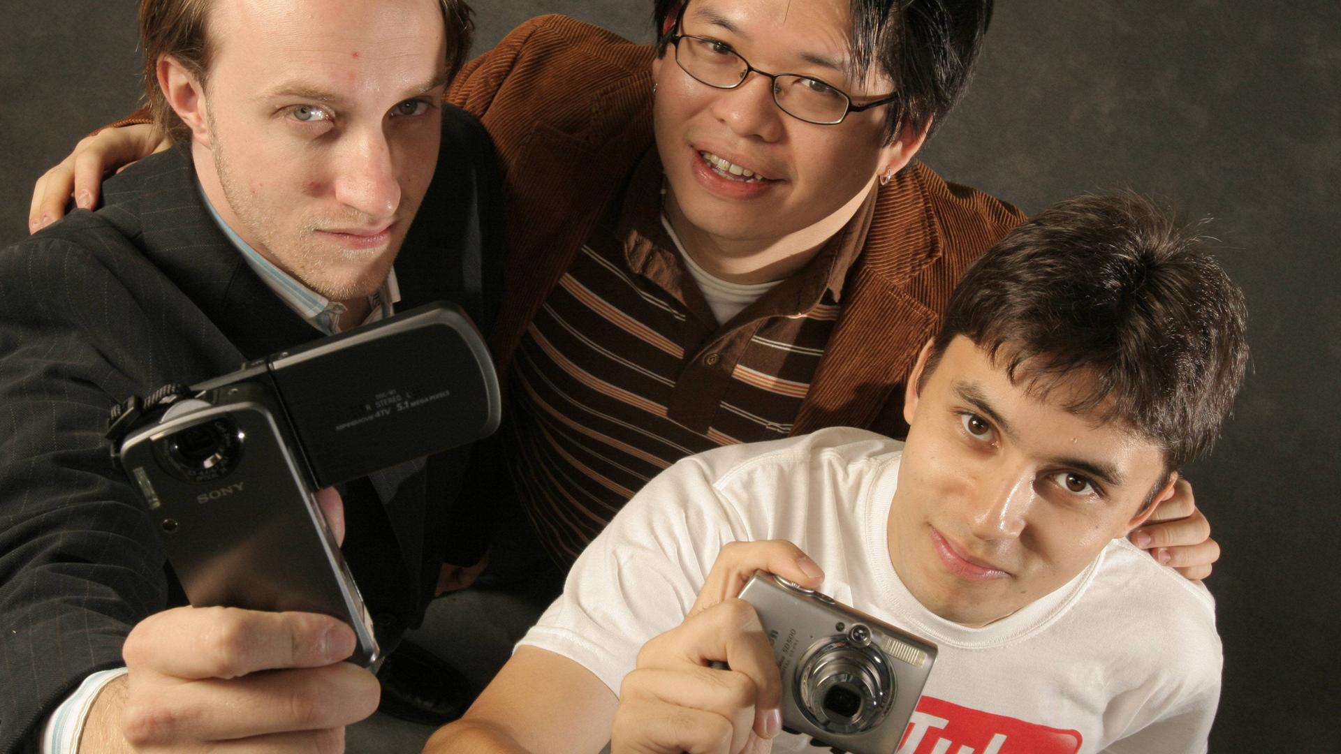 Chad Hurley, Jawed Karim und Steve Chen stehen eng aneinander und schauen nach oben in die Kamera. Einer hält ein Klapphandy in der Hand, der andere eine Kompakt-Digitalkamera. Das Bild ist 20 Jahre alt.