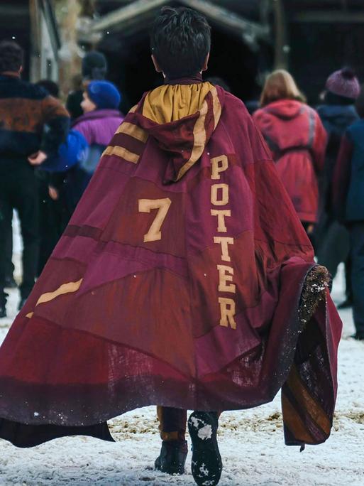 Eine Syene aus der neuen HBO Serie zu Harry Potter. Eine Person mit einem Umhang auf dem die Zahl 7 neben dem Namen Potter steht ist von hinten zu sehen, während sie auf eine Gruppe Personen zugeht.