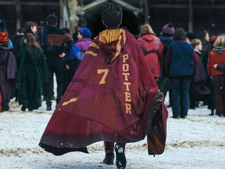 Eine Syene aus der neuen HBO Serie zu Harry Potter. Eine Person mit einem Umhang auf dem die Zahl 7 neben dem Namen Potter steht ist von hinten zu sehen, während sie auf eine Gruppe Personen zugeht.