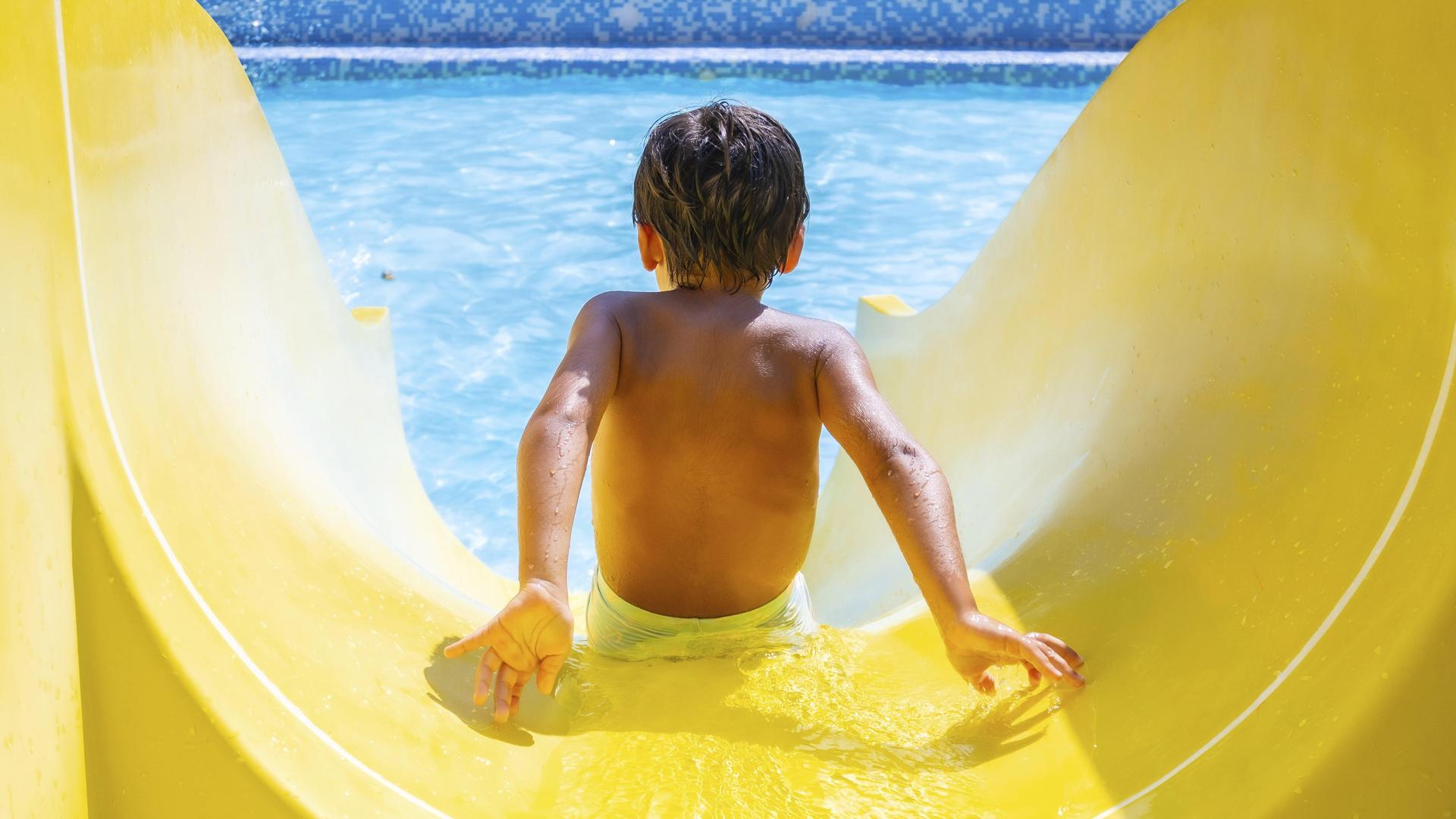 Ein Kind rutscht freudig eine gelbe Wasserrutsche in einem Freibad hinunter.