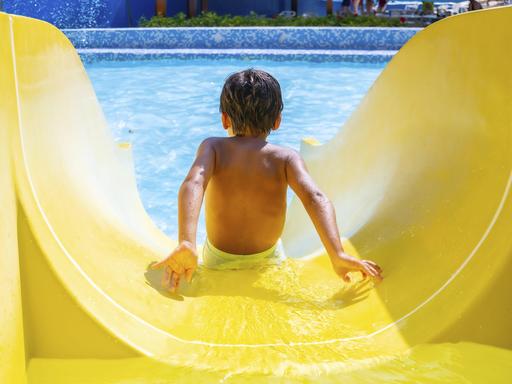 Ein Kind rutscht freudig eine gelbe Wasserrutsche in einem Freibad hinunter.