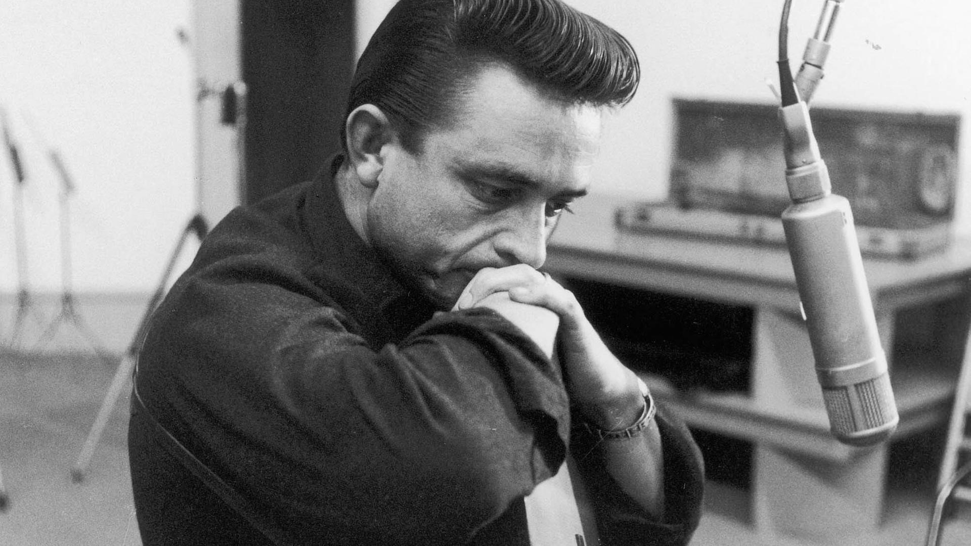 Johnny Cash sitzt mit einer Gitarre nachdenklich vor einem Mikrofon.