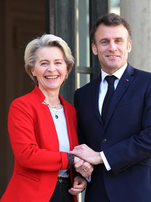 Ursula von der Leyen und Emmanuel Macron stehen in die Kamera lachend nebeneinander. Sie trägt ein rotes Sacko, er ein blaues.