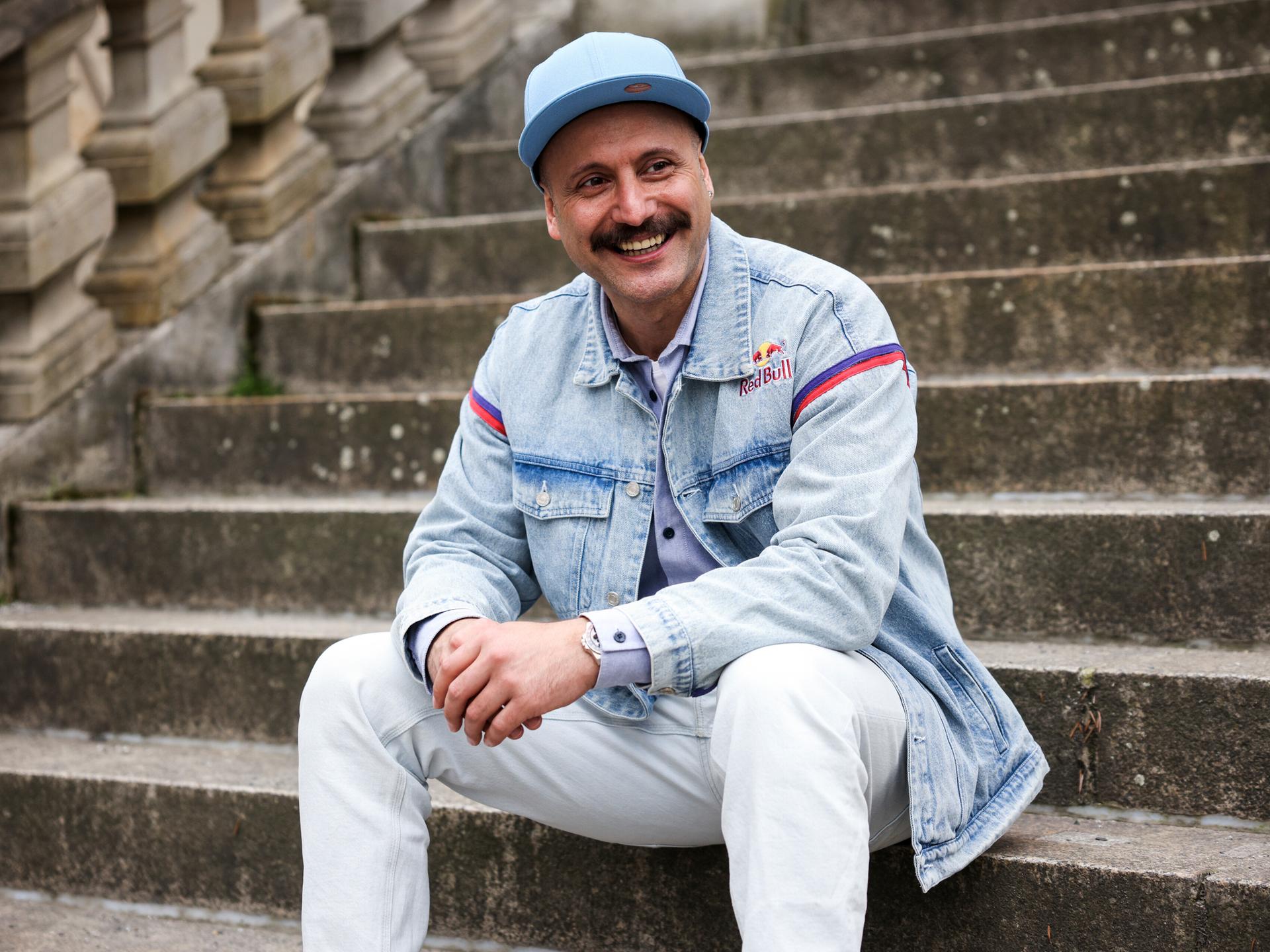 Ein junger Mann mit Schnurrbart und hellblauer Kappe sitzt auf einer steinernen Außentreppe. Er trägt eine helle Jeans sowie eine Jeansjacke und blickt lachend zu seiner linken Seite.