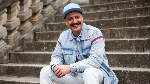 Ein junger Mann mit Schnurrbart und hellblauer Kappe sitzt auf einer steinernen Außentreppe. Er trägt eine helle Jeans sowie eine Jeansjacke und blickt lachend zu seiner linken Seite.