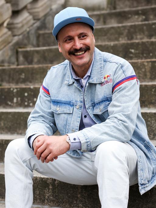 Ein junger Mann mit Schnurrbart und hellblauer Kappe sitzt auf einer steinernen Außentreppe. Er trägt eine helle Jeans sowie eine Jeansjacke und blickt lachend zu seiner linken Seite.