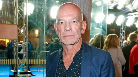 Der damals 57 Jahre alte Martin Wuttke steht in einem Foyer und schaut in die Kamera. Er hat eine Glatze und trägt ein blaues Jacket, darunter ein dunkleres, gemustertes, weit geöffnetes Hemd.