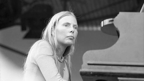 Joni Mitchell bei Konzert auf der britischen Isle of Wight im August 1970 mit geschätzten 700.000 Besuchern. Die Künstlerin sitzt auf dem Schwarz-Weiß-Bild vor dem Klavier und schaut zurückhaltend Richtung Publikum. 