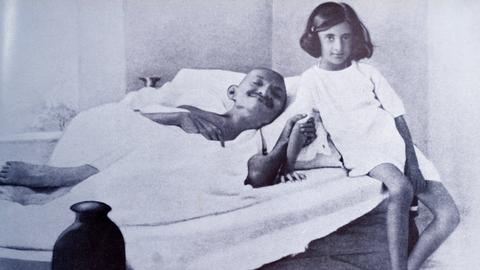Ein Mann, Mahatma Gandhi liegt kräftelos in einem Bett. Er hungert. Neben ihm sitzt ein junges Mädchen, das in die Kamera lächelt. Ein Mann, Mahatma Gandhi liegt kräftelos in einem Bett. Er hungert. Neben ihm sitzt ein junges Mädchen, das in die Kamera lächelt.
