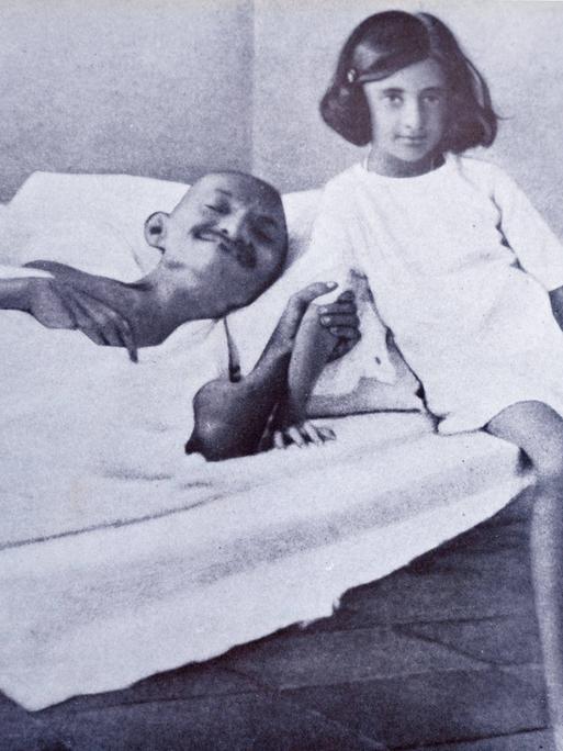 Ein Mann, Mahatma Gandhi liegt kräftelos in einem Bett. Er hungert. Neben ihm sitzt ein junges Mädchen, das in die Kamera lächelt. 
