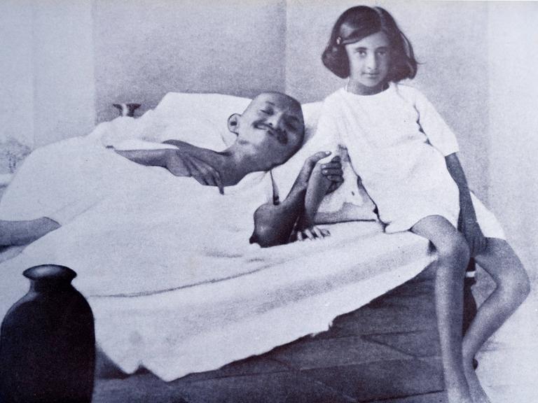 Ein Mann, Mahatma Gandhi liegt kräftelos in einem Bett. Er hungert. Neben ihm sitzt ein junges Mädchen, das in die Kamera lächelt. 