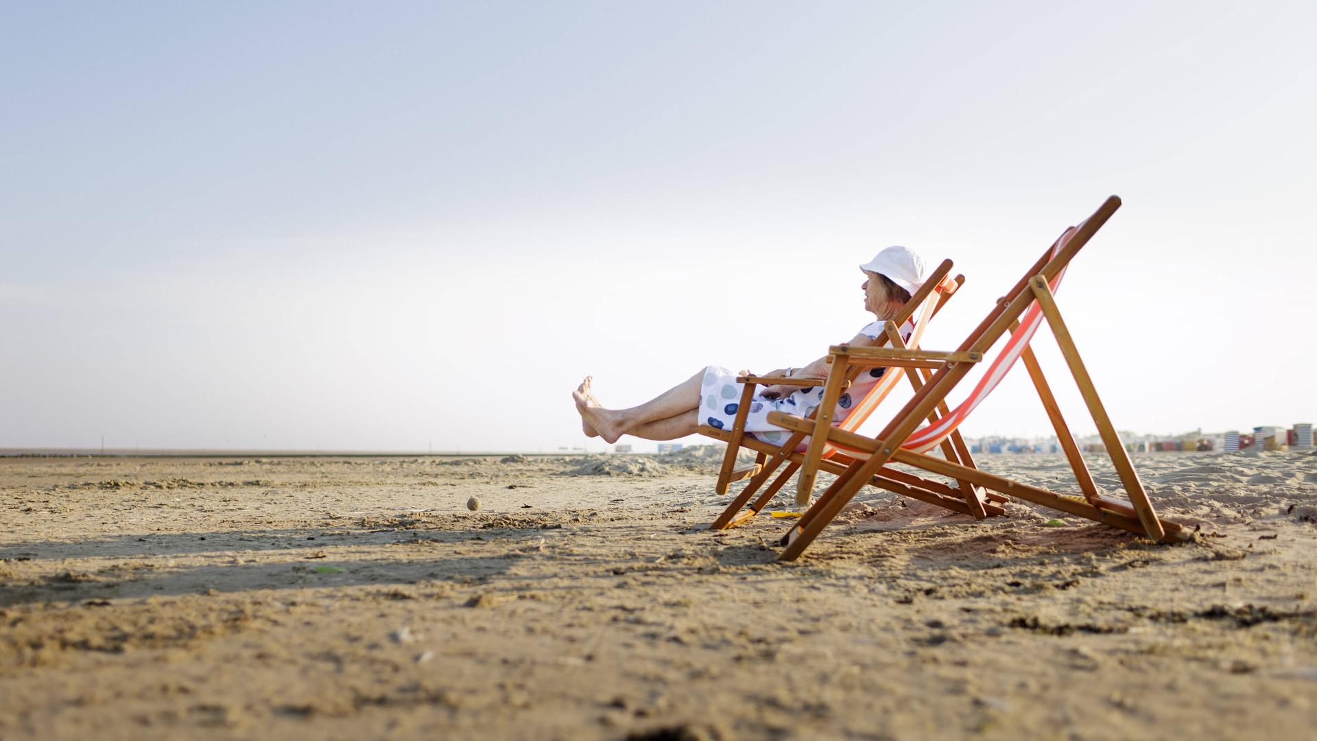 Eine Frau sitzt in einem Strandstuhl. Sie trägt ein Kleidung und einen Sonnenhut.