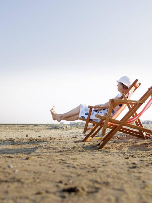 Eine Frau sitzt in einem Strandstuhl. Sie trägt ein Kleidung und einen Sonnenhut.