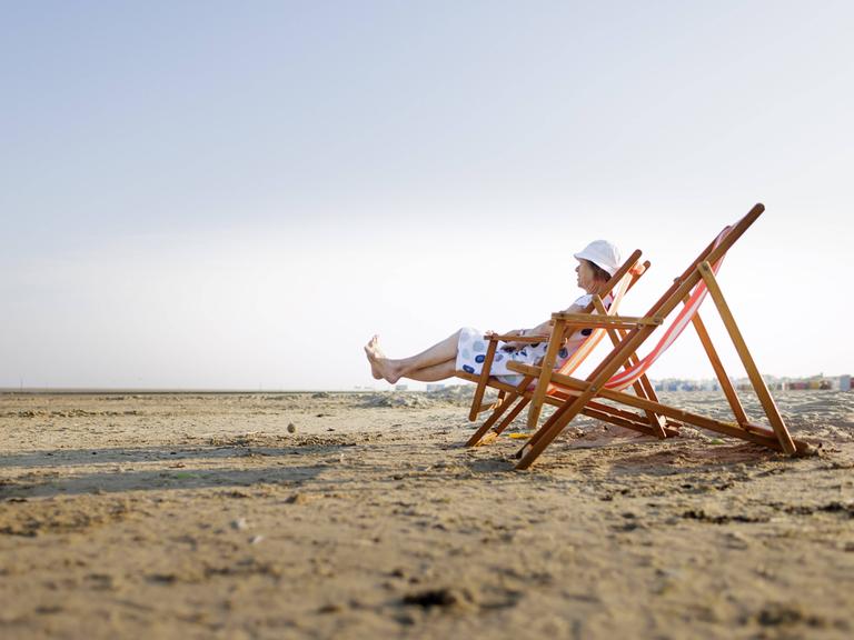 Eine Frau sitzt in einem Strandstuhl. Sie trägt ein Kleidung und einen Sonnenhut.