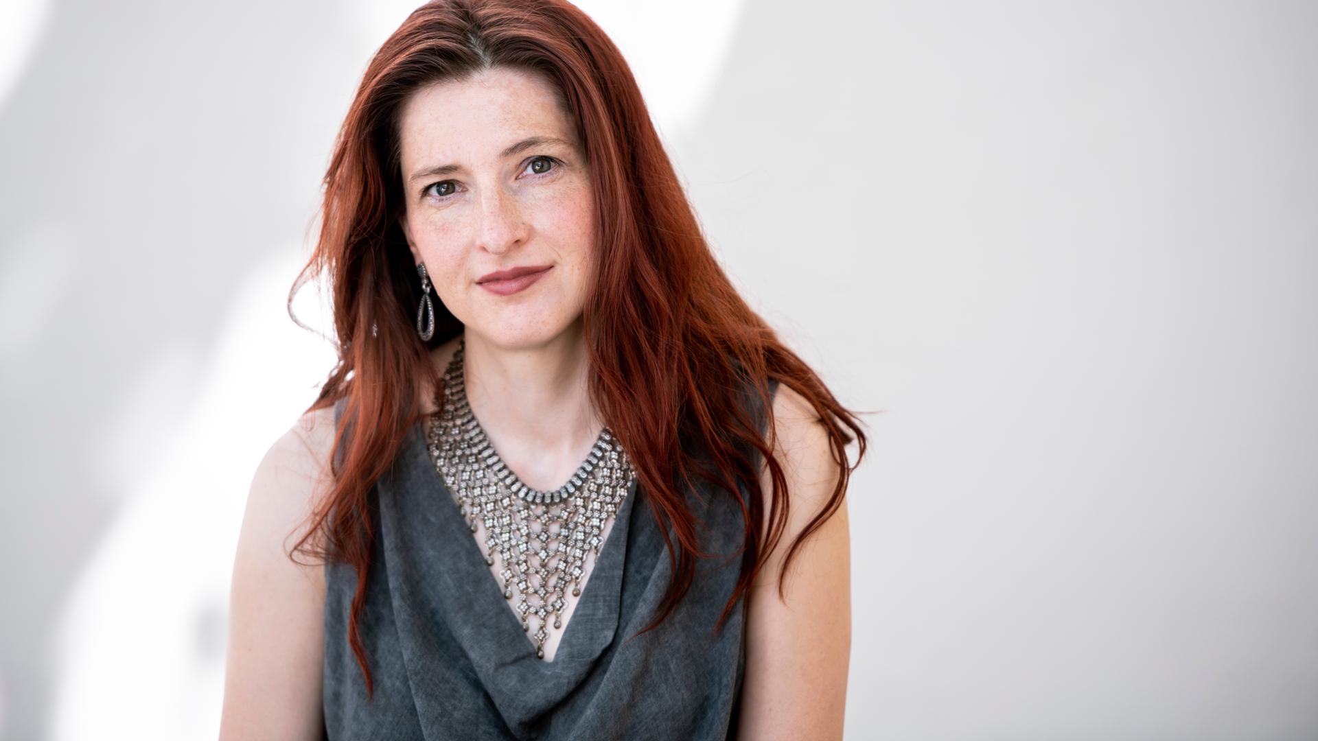 Daniela Danz schaut in die Kamera. Sie hat langes rotbraunes Haar und trägt ein graues Oberteil mit Wasserfallausschnitt zu auffälligem Schmuck. Daniela Danz schaut in die Kamera. Sie hat langes rotbraunes Haar und trägt ein graues Oberteil mit Wasserfallausschnitt zu auffälligem Schmuck.