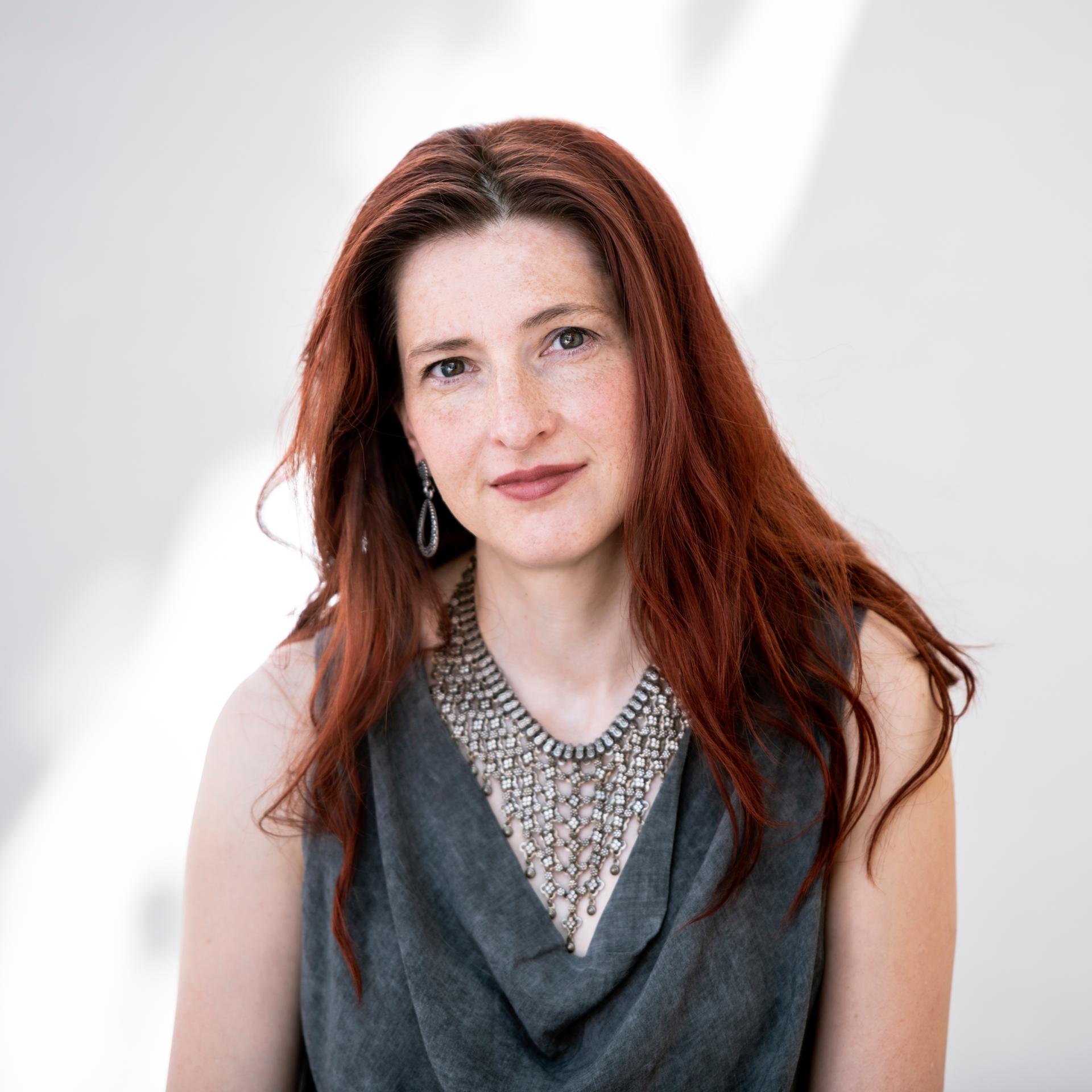 Daniela Danz schaut in die Kamera. Sie hat langes rotbraunes Haar und trägt ein graues Oberteil mit Wasserfallausschnitt zu auffälligem Schmuck.