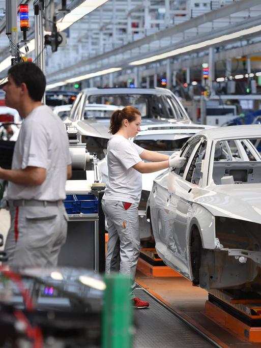 Eine Produktionsanlage von Audi. An den Karosserien arbeiten eine Frau und zwei Männer.