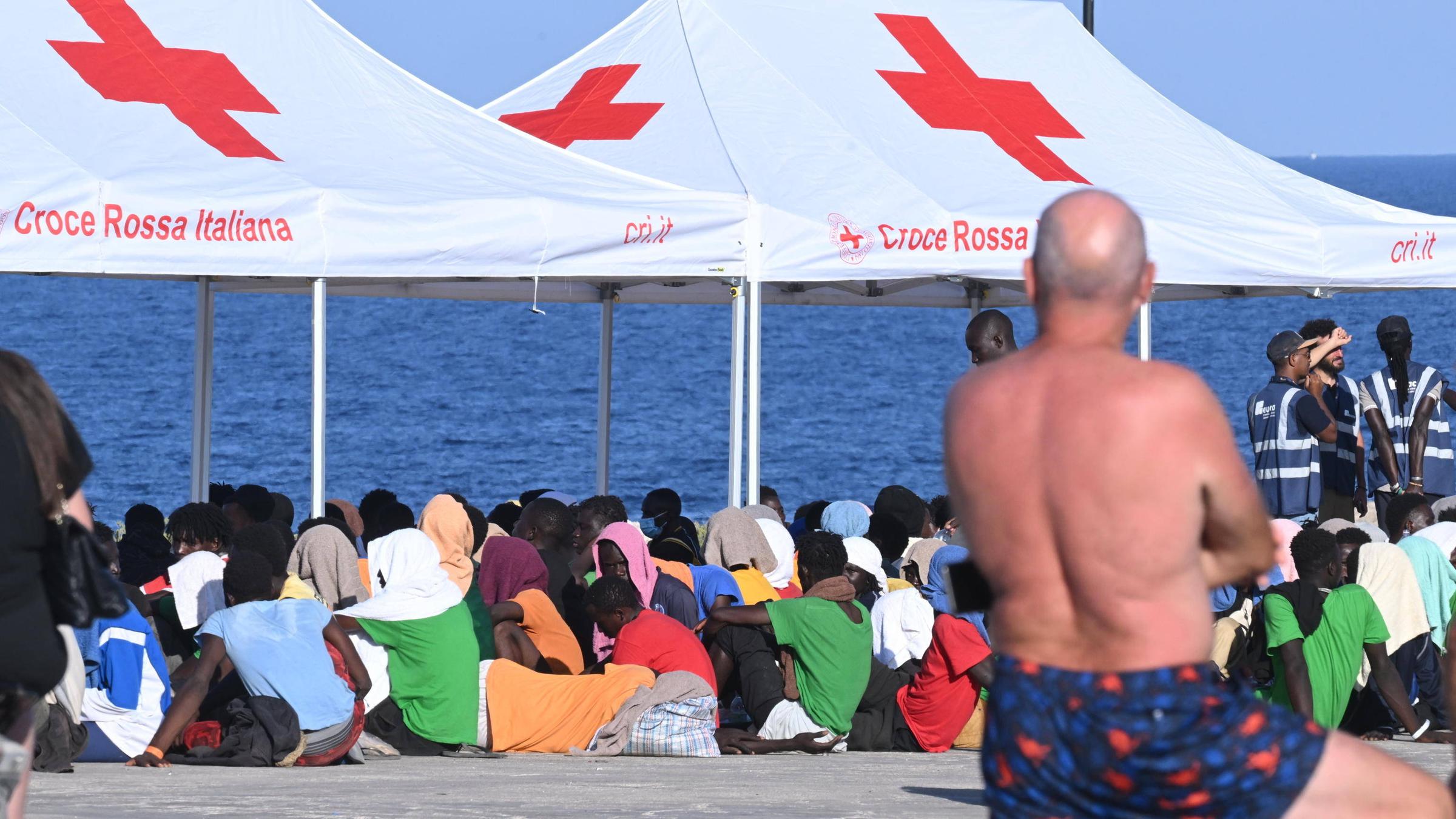 Geflüchtete sitzen unter einem Zelt des Roten Kreuzes im italienischen Lampedusa. Im Vordergrund steht ein Mann in Badehose und beobachtet sie. 