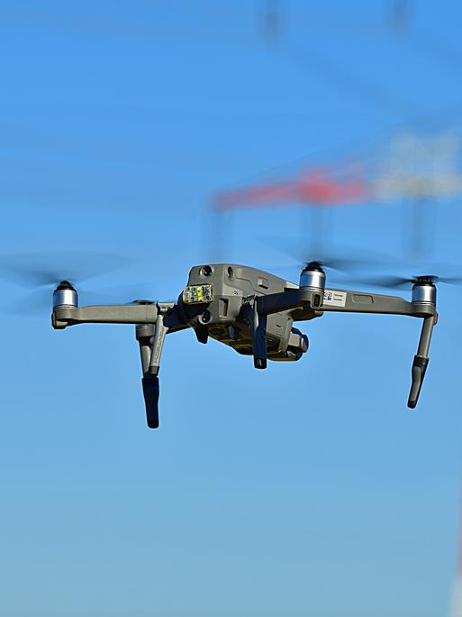 Eine schwarze Drohne fliegt vor einem rot-weißen Strommasten.