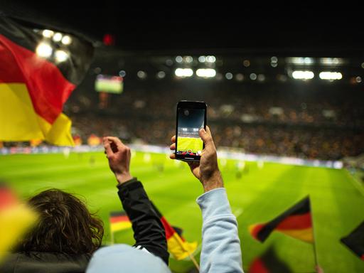 In einem Fußballstadion werden Deutschland-Flaggen geschwenk, jemand hält ein Smartphone in die Höhe