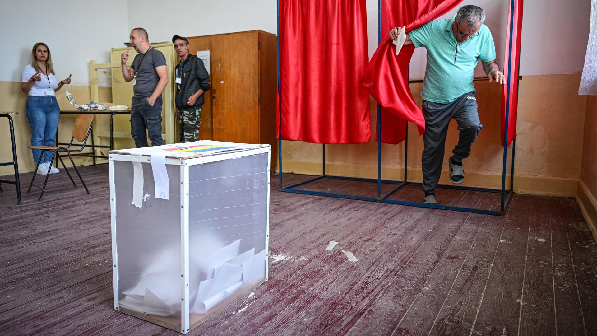 Ein Mann tritt aus einer Wahlkabine hinter einem roten Vorhang hervor. Die Wahlurne steht unmittelbar vor ihm im Raum.