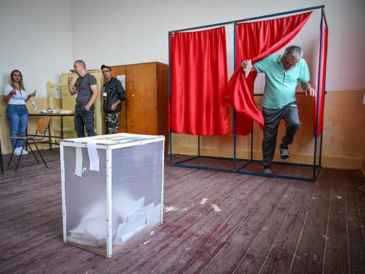 Ein Mann tritt aus einer Wahlkabine hinter einem roten Vorhang hervor. Die Wahlurne steht unmittelbar vor ihm im Raum.