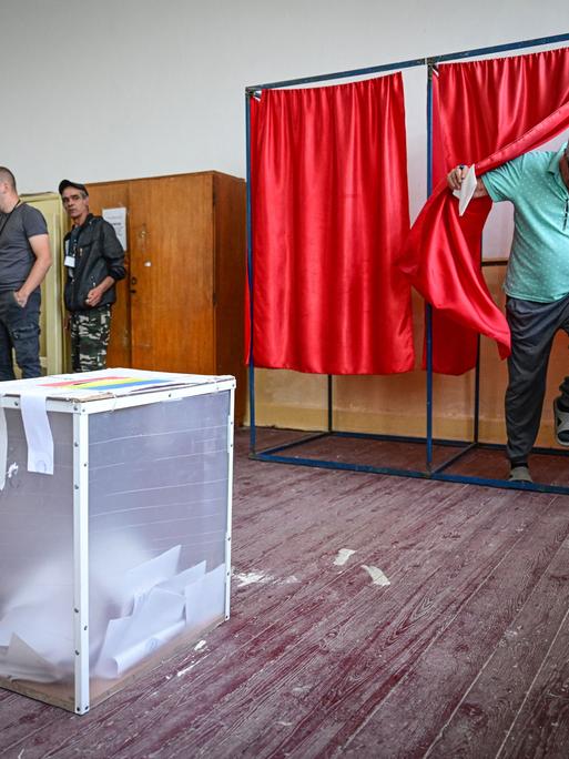 Ein Mann tritt aus einer Wahlkabine hinter einem roten Vorhang hervor. Die Wahlurne steht unmittelbar vor ihm im Raum.