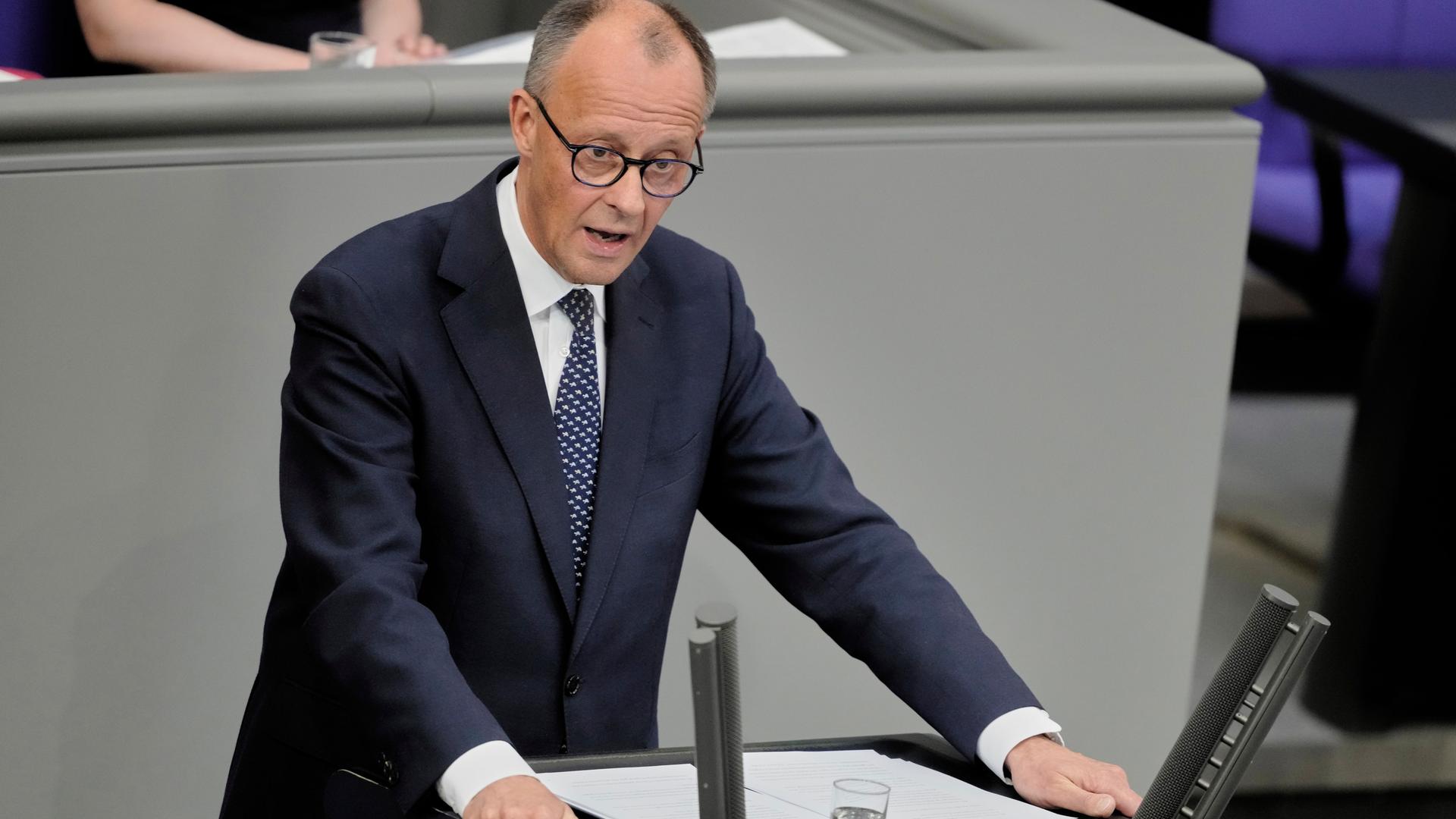 Das Foto zeigt Friedrich Merz am Rednerpult des Bundestags. Er trägt dunkelblauen Anzug und eine blaue Krawatte. Seine Arme stützt er auf das Pult. 