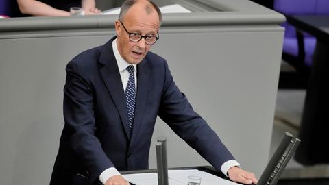 Das Foto zeigt Friedrich Merz am Rednerpult des Bundestags. Er trägt dunkelblauen Anzug und eine blaue Krawatte. Seine Arme stützt er auf das Pult. 