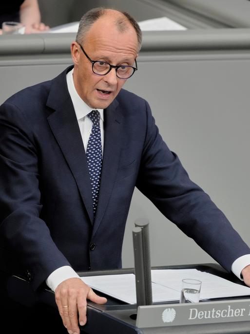 Das Foto zeigt Friedrich Merz am Rednerpult des Bundestags. Er trägt dunkelblauen Anzug und eine blaue Krawatte. Seine Arme stützt er auf das Pult. 