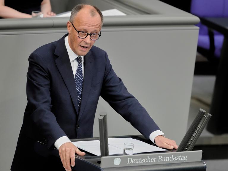 Das Foto zeigt Friedrich Merz am Rednerpult des Bundestags. Er trägt dunkelblauen Anzug und eine blaue Krawatte. Seine Arme stützt er auf das Pult. 