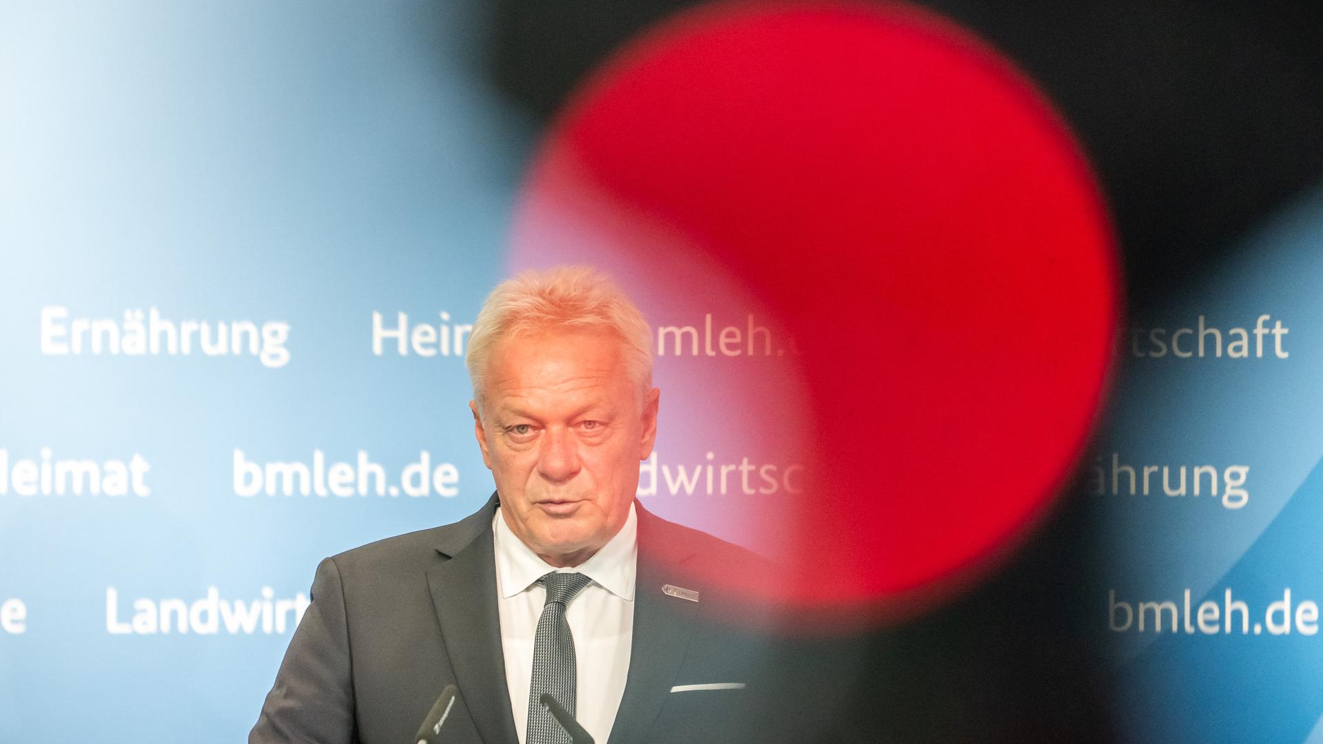 Zu sehen ist Alois Rainer an einem Redepult mit Mikrofonen vor einer blauen Wand. Durch eine Spiegelung in der Linse des Fotoapparates ist ein großer roter Kreis zu sehen.