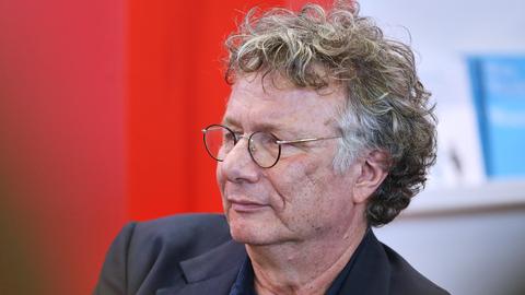Der Schriftsteller Ingo Schulze von der Seite im Porträt. Er trägt eine Brille und ist vor einem roten Hintergrund im dunklen Jackett. 