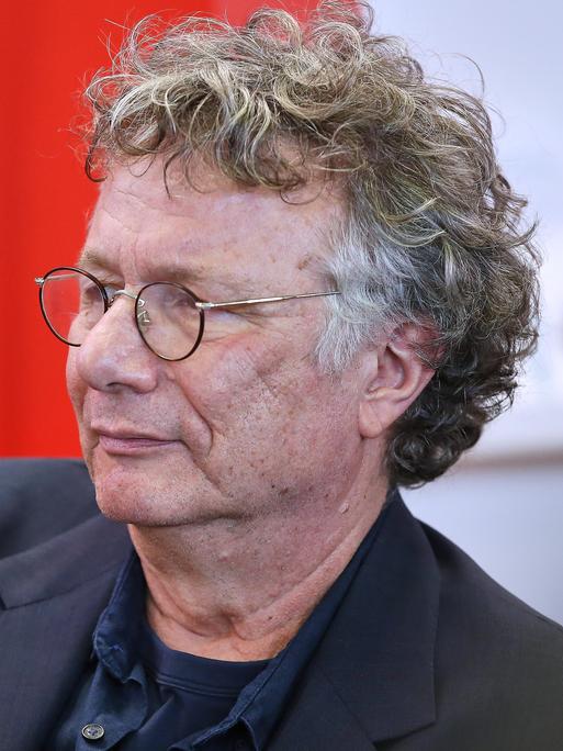Der Schriftsteller Ingo Schulze von der Seite im Porträt. Er trägt eine Brille und ist vor einem roten Hintergrund im dunklen Jackett. 