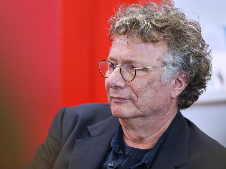 Der Schriftsteller Ingo Schulze von der Seite im Porträt. Er trägt eine Brille und ist vor einem roten Hintergrund im dunklen Jackett. 