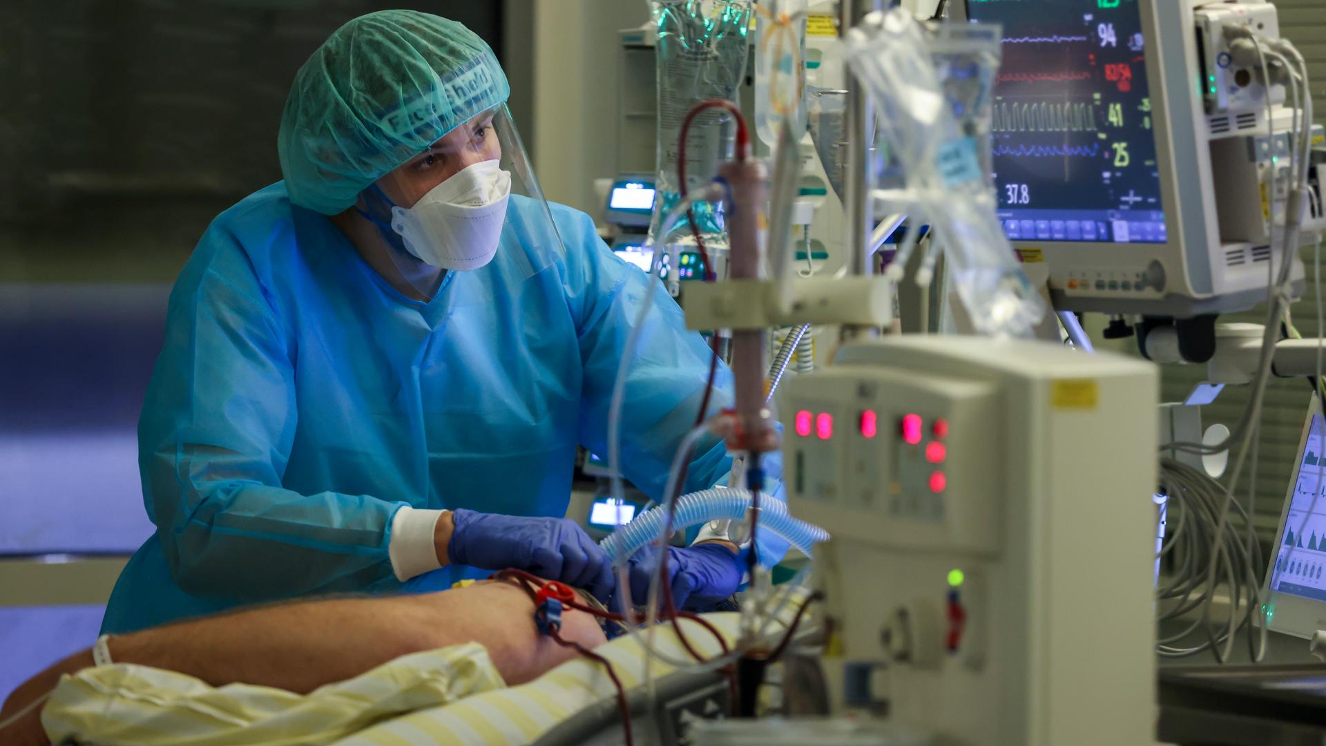 Ein Intensivpfleger versorgt einen Covid-19-Patienten auf einer Intensivstation. Um das Bett herum stehen mehrere medizinie Apparate. Der Pfleger schaut konzentriert auf einen Bildschirm.