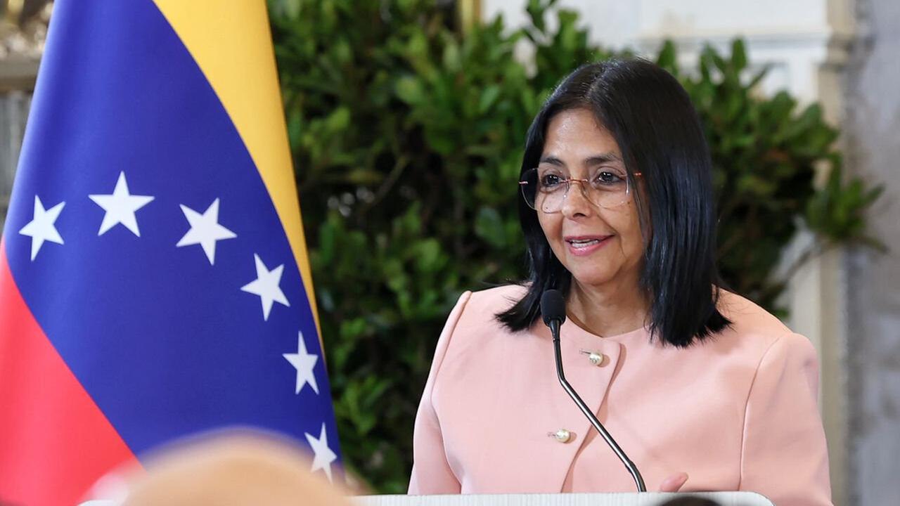 Delcy Rodríguez steht an einem Redepult. Sie trägt einen rosafarbenen Blazer. Neben ihr steht die Nationalflagge Venezuelas.
