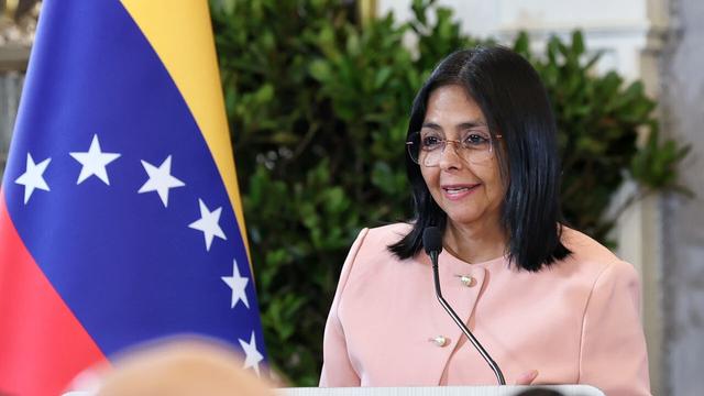 Delcy Rodríguez steht an einem Redepult. Sie trägt einen rosafarbenen Blazer. Neben ihr steht die Nationalflagge Venezuelas.