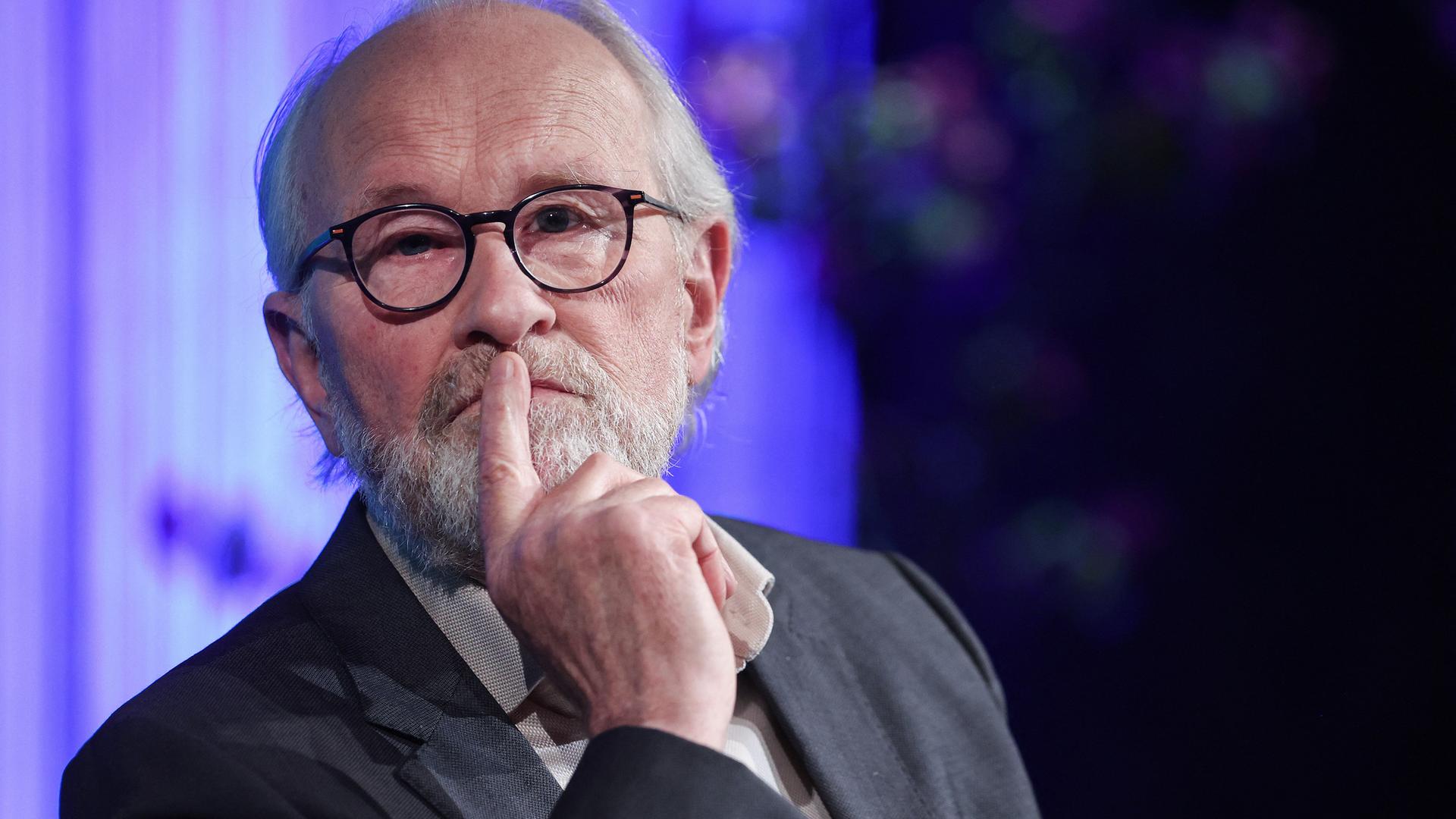 Lukas Beckmann sitzt nachdenklich auf einem Podium und schaut leicht an der Kamera vorbei. Er trägt eine schwarze Brille und einen dunkelgrauen Anzug.