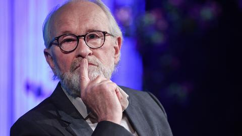 Lukas Beckmann sitzt nachdenklich auf einem Podium und schaut leicht an der Kamera vorbei. Er trägt eine schwarze Brille und einen dunkelgrauen Anzug.