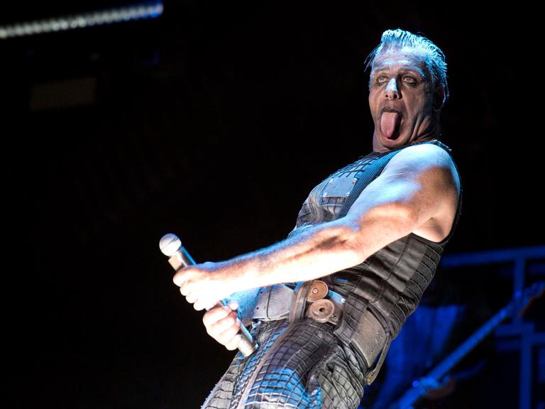 Der umstrittene Rammstein-Frontmann Till Lindemann steht geschminkt auf einer Bühne und macht mit dem Mikrofon eine Geste, die an einen Phallus erinnert. 