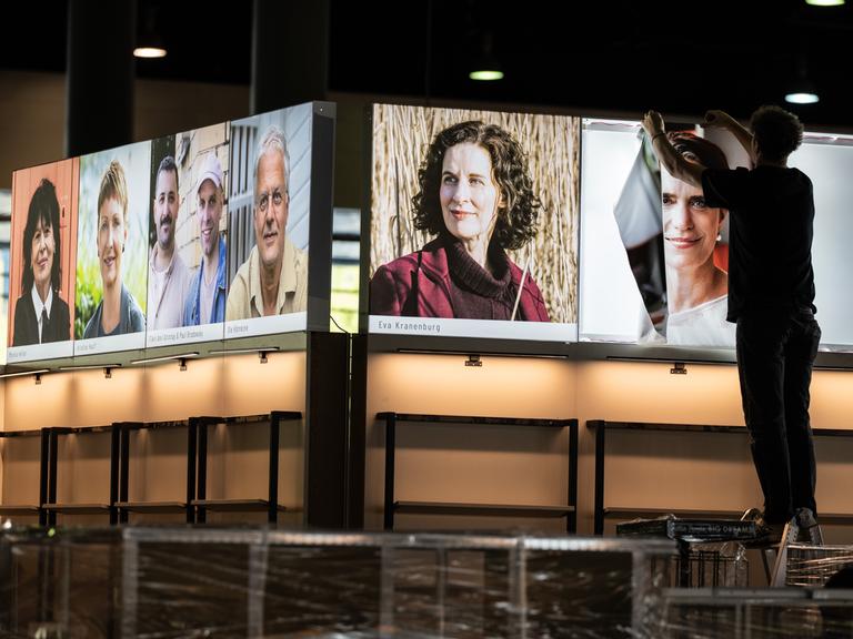 Messebauer errichten den Stand eines Buchverlages auf der Buchmesse und hängen Fotos von Autorinnen und Autoren auf.