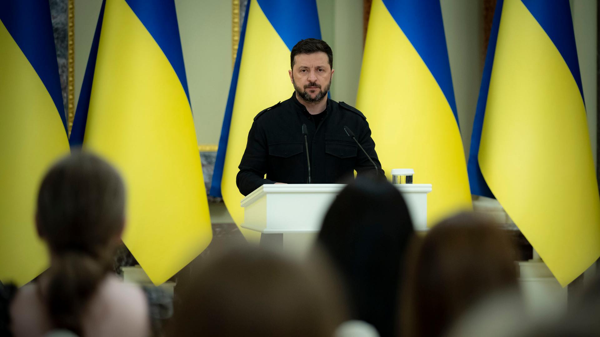 Der ukrainische Präsident Wolodymyr  Selenskyj steht am Mikrofon, hinter ihm stehen ukrainische Flaggen in einer langen Reihe
