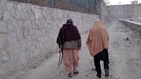 Zwei Männer laufen an einer hohen Mauer mit Stacheldraht, einer trägt ein Gewehr unter dem Arm, Afghanistan 2022.