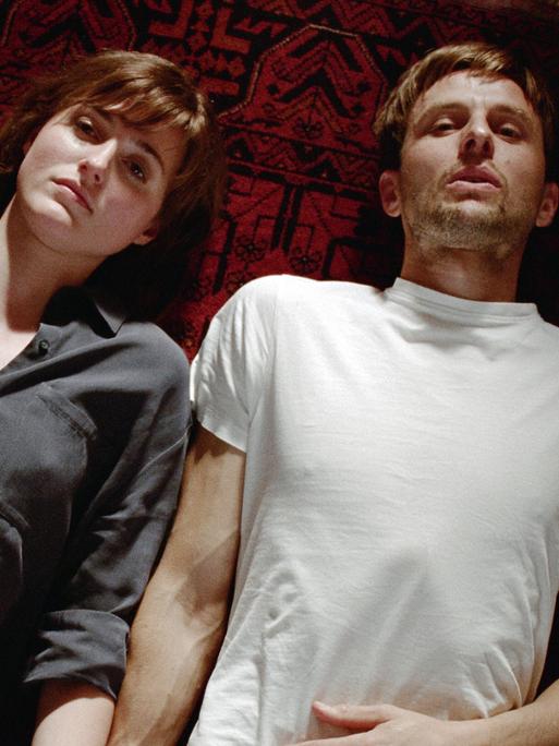 Zwei Menschen liegen auf einem Teppich. Links eine Frau mit dunklen Haaren und einem grauen Hemd, rechts ein Mann mit weißem T-Shirt. Zwei Menschen liegen auf einem Teppich. Links eine Frau mit dunklen Haaren und einem grauen Hemd, rechts ein Mann mit weißem T-Shirt.