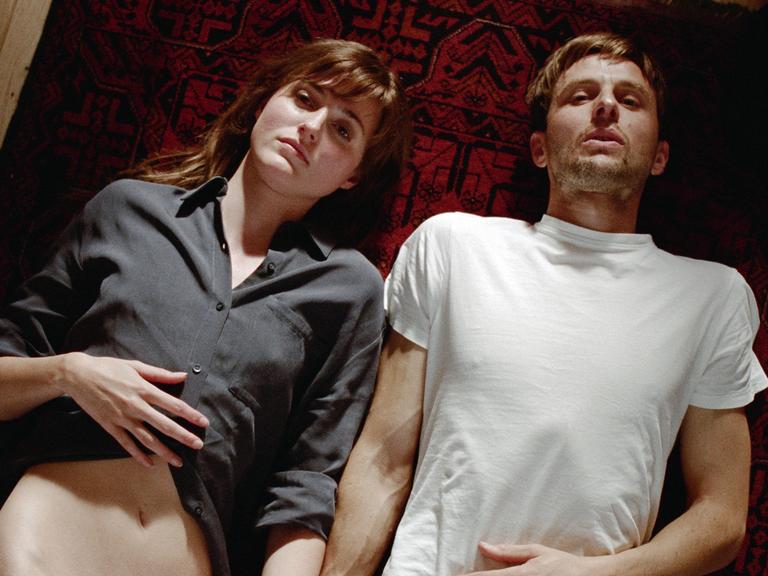 Zwei Menschen liegen auf einem Teppich. Links eine Frau mit dunklen Haaren und einem grauen Hemd, rechts ein Mann mit weißem T-Shirt. Zwei Menschen liegen auf einem Teppich. Links eine Frau mit dunklen Haaren und einem grauen Hemd, rechts ein Mann mit weißem T-Shirt.