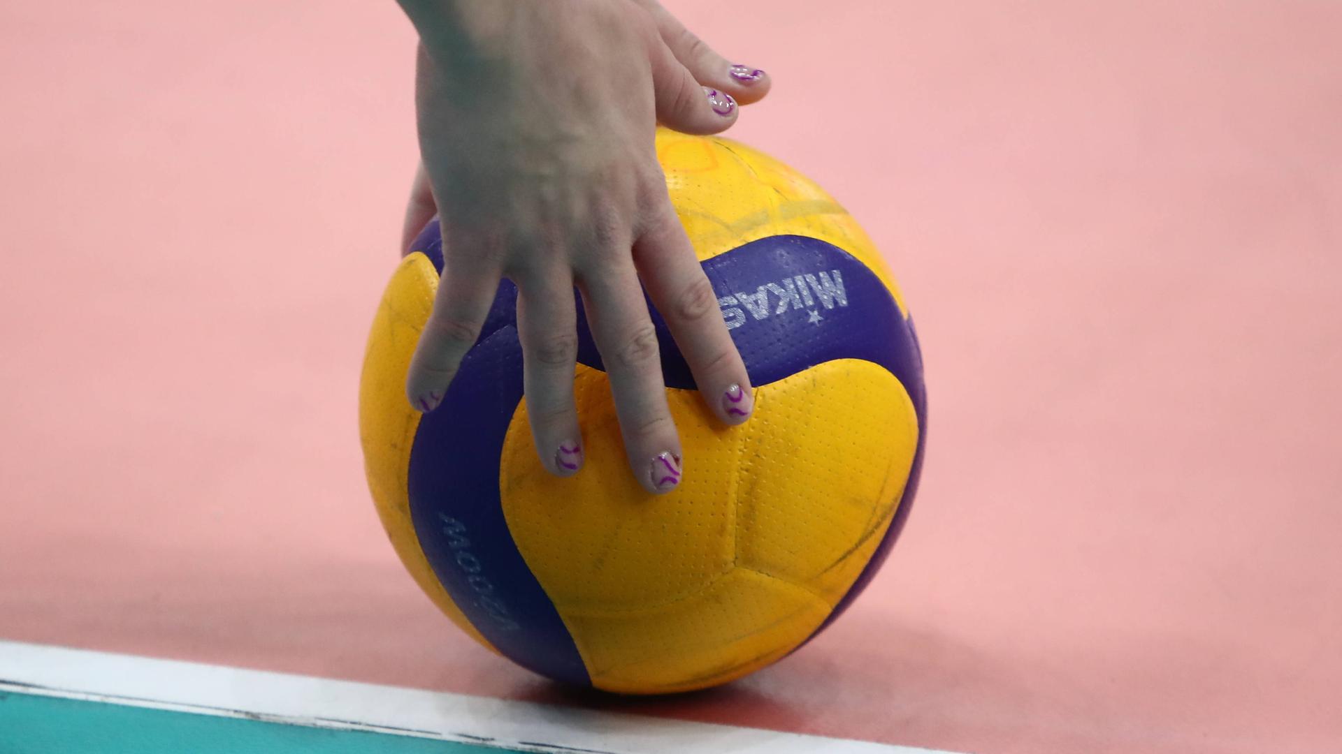 Hände mit lackierten Fingernägel greifen nach einem zweifarbigen Volleyball, der auf dem Hallenboden liegt.